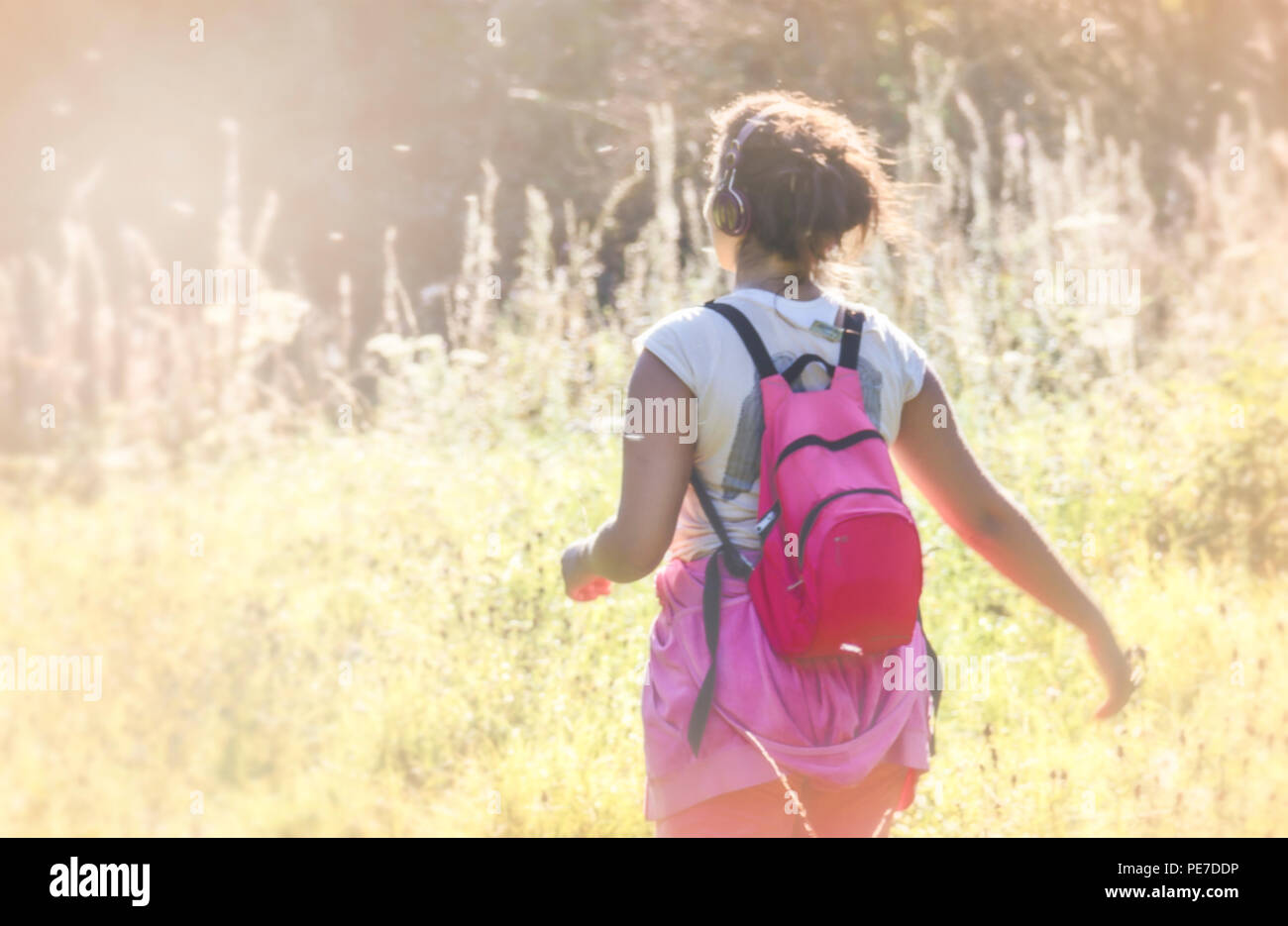 A young woman with a pink backpack and wearing headphones strolling through a hazy summer meadow during the heatwave Stock Photo