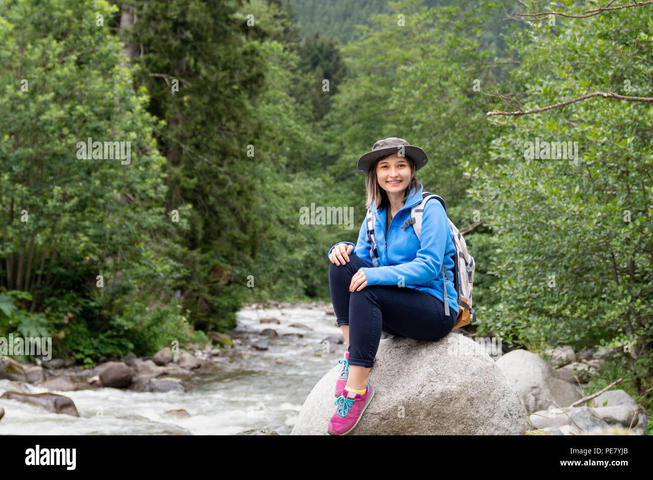 Woman with backpack sitting on the rock and smiling on a background of forest. Stock Photo