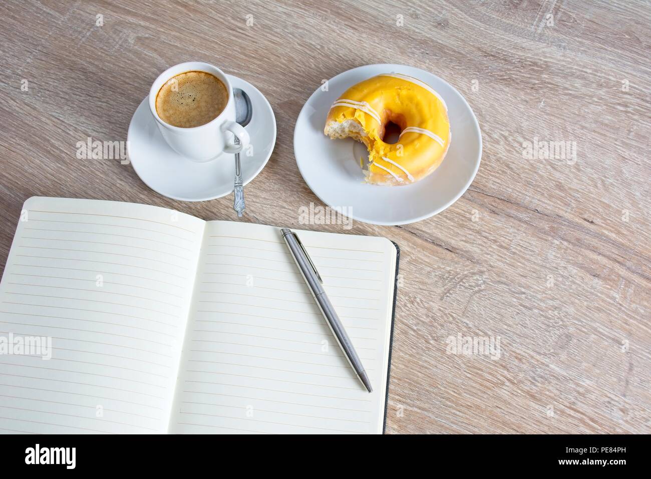 Note book and pen, doughnut on a plate, small cup of coffee, jug of milk and sugar pot on table top. Stock Photo