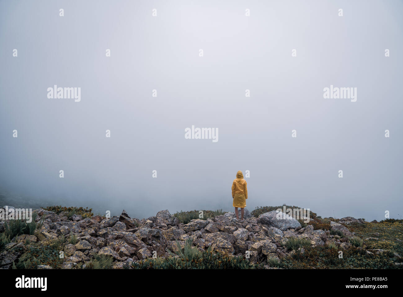 Female in yellow raincoat, jeans shorts standing at top of mountain with view of peaks at horizon. Landscape. Nature. Valley. Travel. Freedom. Vacation. Hills. Success. Contemplation. Flight Stock Photo