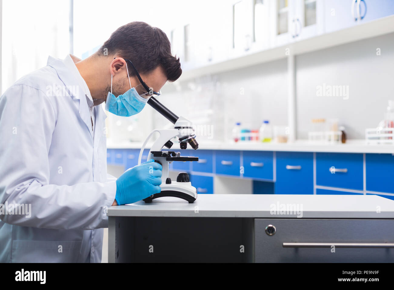 Successful study. Skillful ambitious male lab assistant standing in profile while scrutinizing sample and using microscope Stock Photo