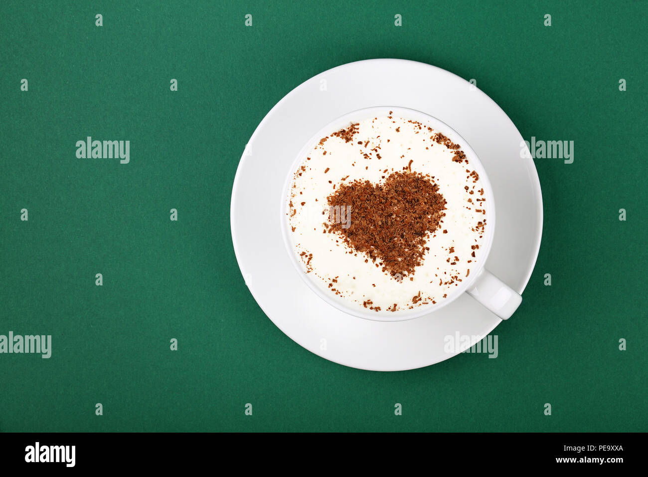 Close up one full white cup of frothy latte cappuccino coffee with heart shaped brown chocolate art, on saucer on green table background, elevated top Stock Photo