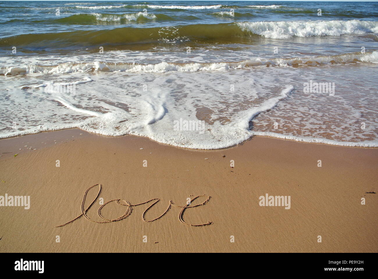 A word 'love' handwritten in a cursive style on the red sand on a beach with the blue sea and a wave in the upper part, Prince Edward Island Stock Photo