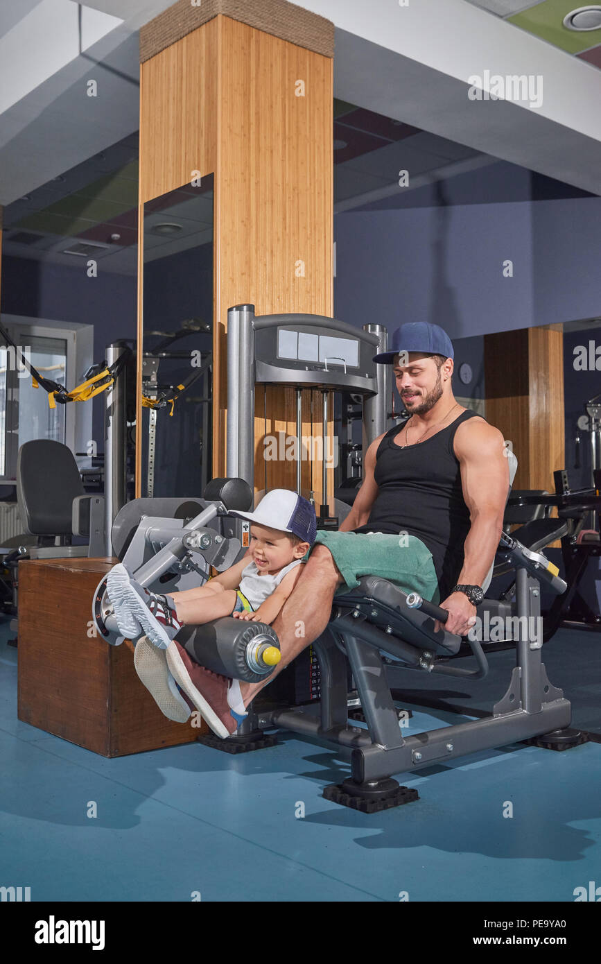 Funny training of father and son in gym with heavy simulators. Sporty man keeping child on knees lifting metallic dumbbells. Wearing shorts, cap, singlet. Spending good, family time together. Stock Photo
