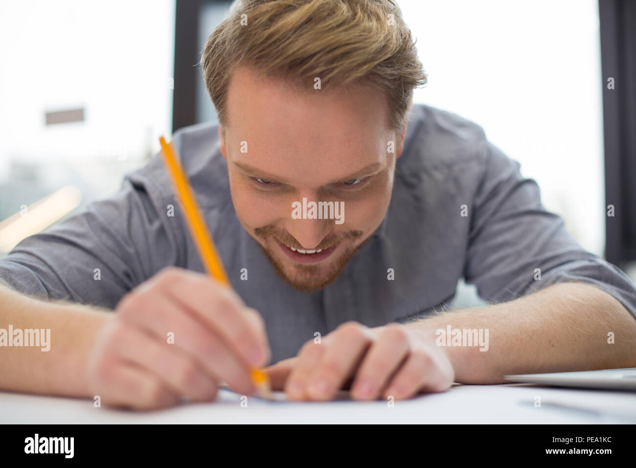 Pleasurable job. Joyful nice delighted man sitting at the table and doing a drawing while concentrating on his work Stock Photo