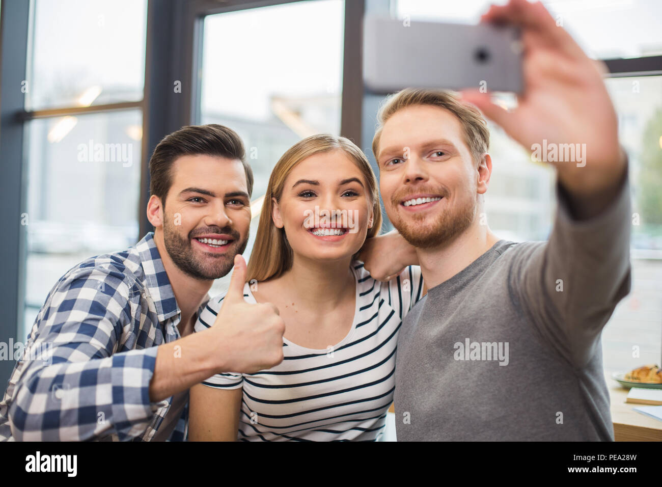 And now smile. Nice pleasant delighted man standing together with his friends and holding a smartphone while taking selfie Stock Photo