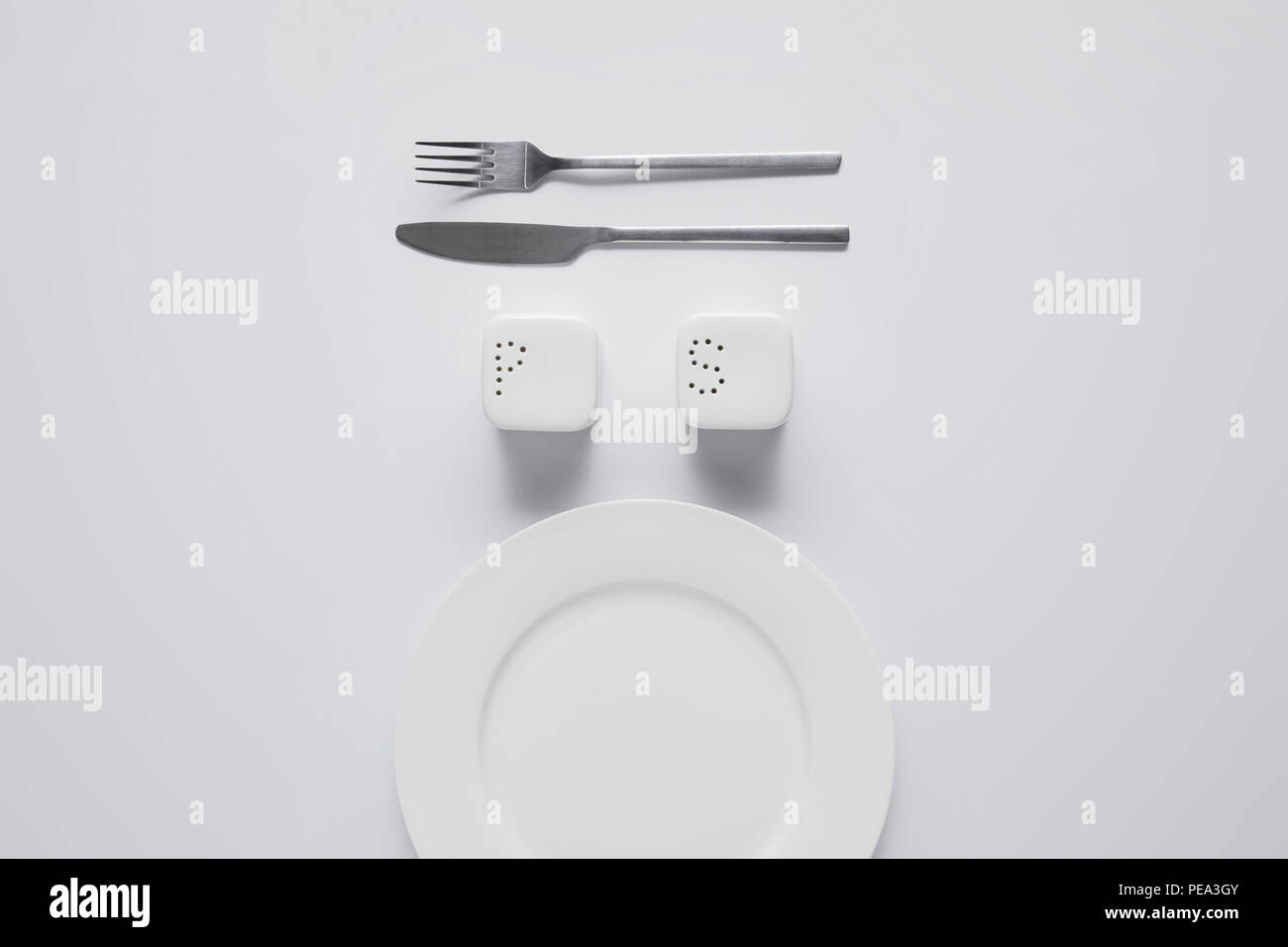 top view of arranged plate, fork, knife, saltcellar and pepper caster on white table, minimalistic concept Stock Photo