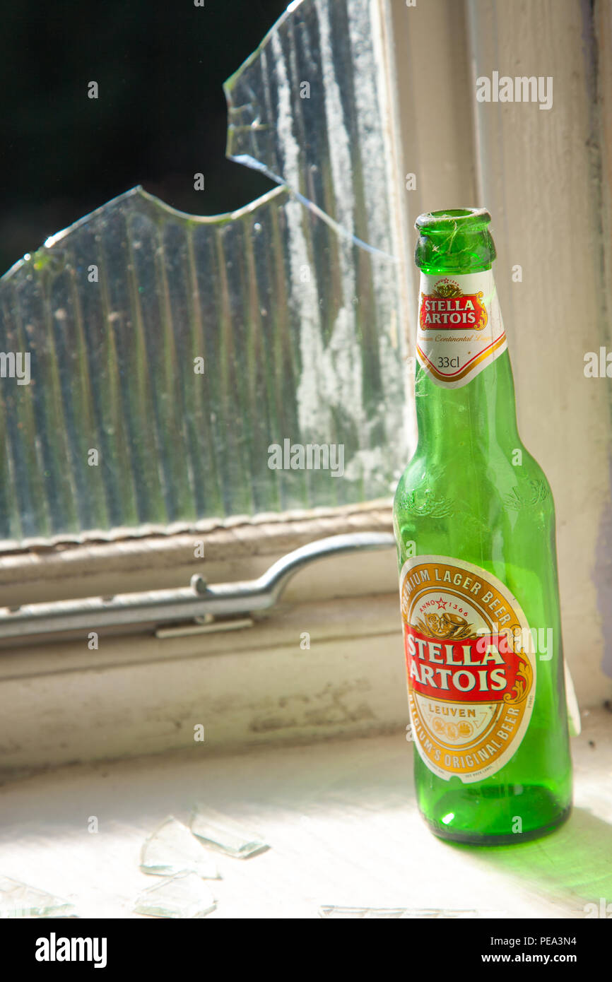 A bottle of Stella Artois beer sits on a window sill by a broken window Stock Photo
