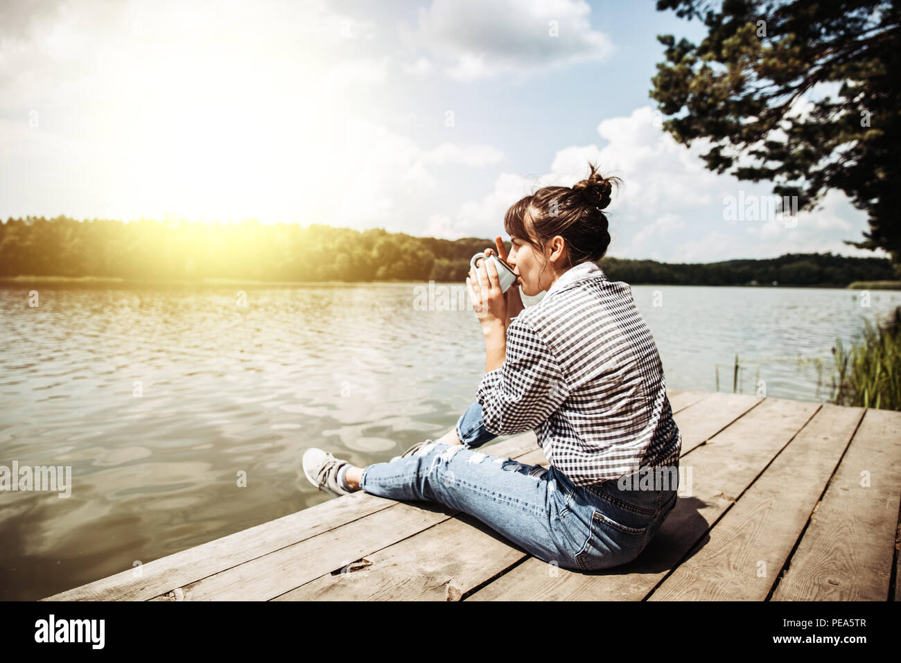 young adult woman sitting on wooden dock drinking coffee and looking at ...
