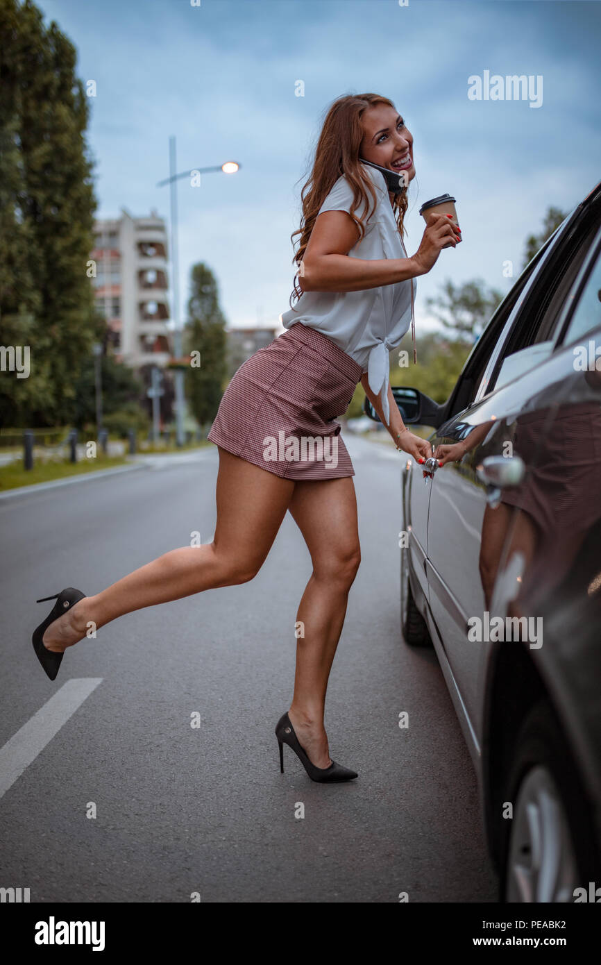 A young beautiful woman in a short skirt is talking on the smartphone,  holding a cup of coffee and is going to enter in the car Stock Photo - Alamy