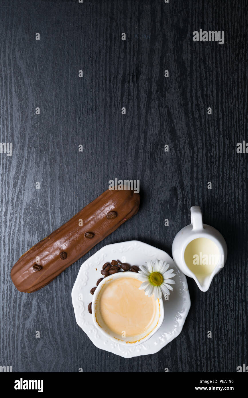 wooden plate with Coffe eclairs with cup of fresh white coffee, pitcher of cream, and coffee beans on Top view. Stock Photo