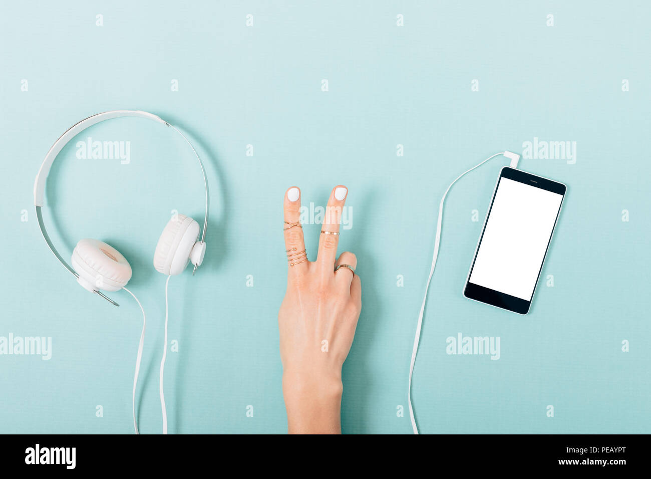 Woman's hand making victory gesture near headphones connected to smart phone on blue background, flat lay. Top view blank screen for the music app des Stock Photo