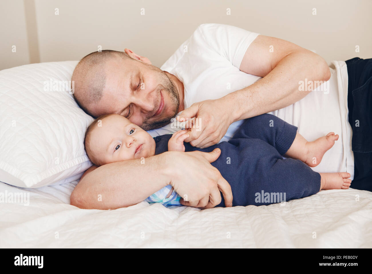 Portrait of middle age Caucasian father in white t-shirt lying in bed with newborn baby son holding his hands fingers, parenting childhood bonding con Stock Photo