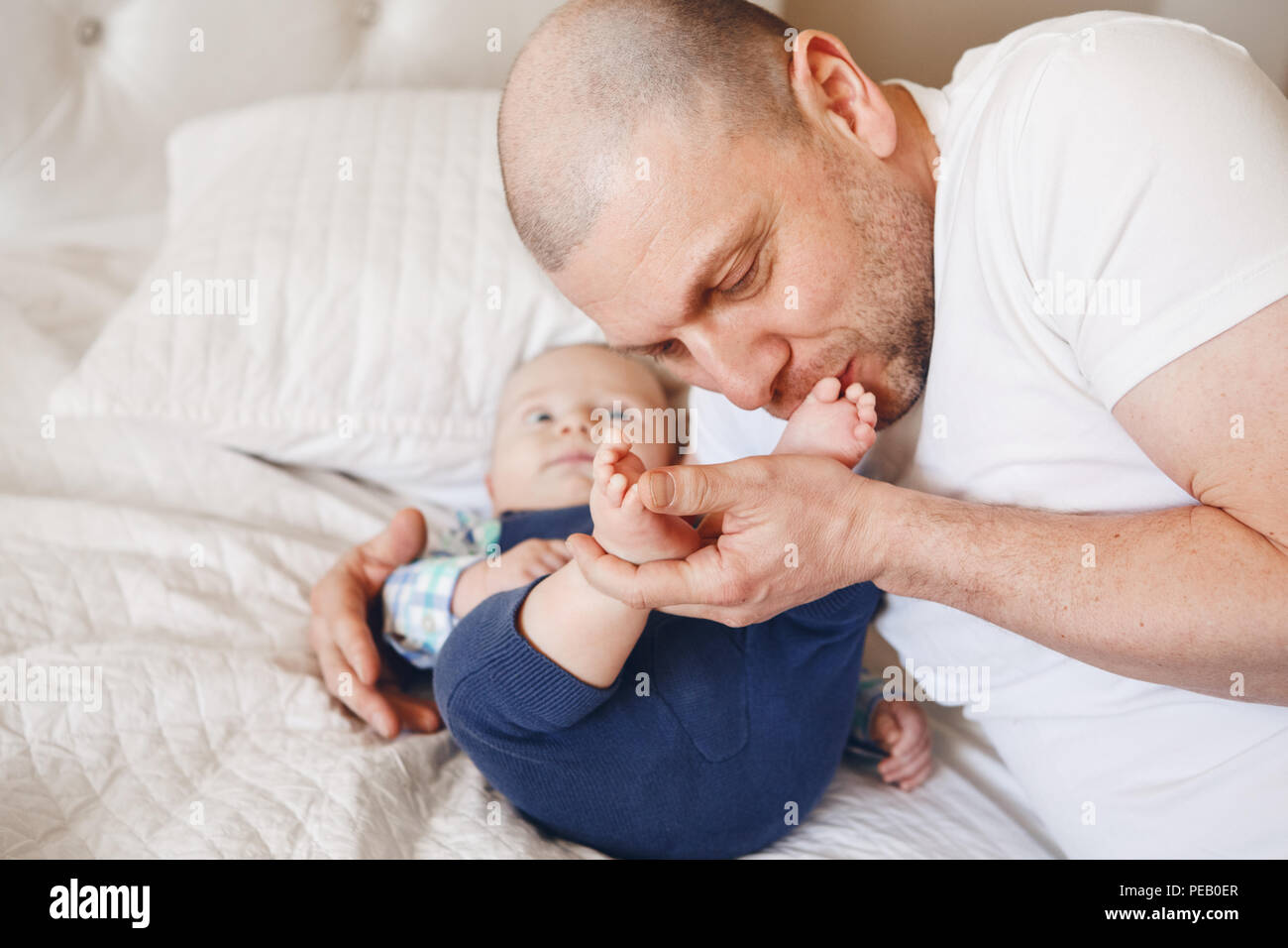 Touching portrait of middle age Caucasian father in white t-shirt lying in bed with newborn baby son kissing his feet toes, parenting childhood bondin Stock Photo