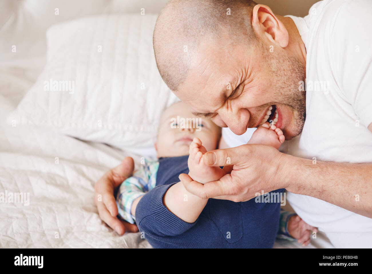 Funny portrait of middle age Caucasian father in white t-shirt lying in bed with newborn baby son kissing biting his feet toes, parenting childhood bo Stock Photo