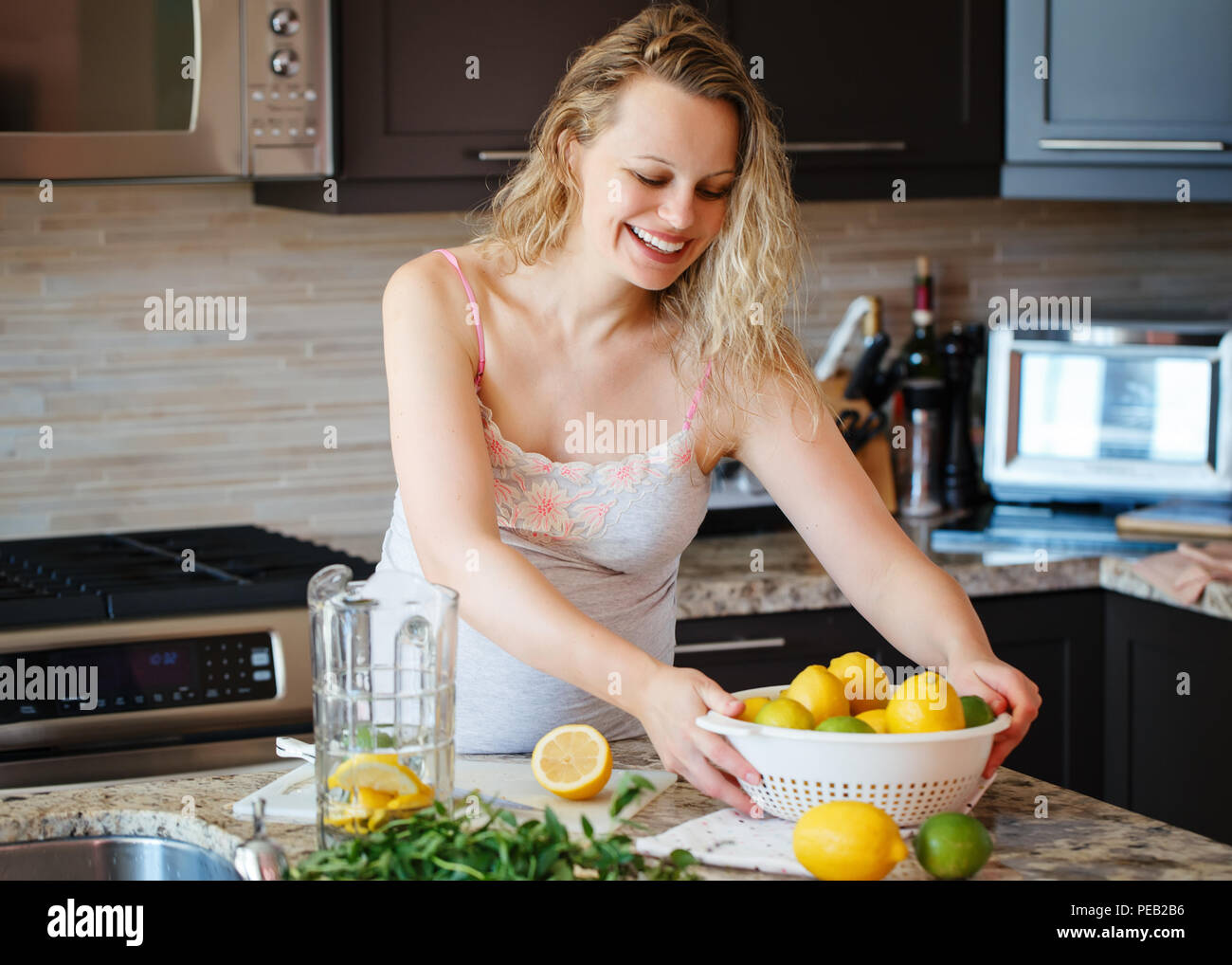 Portrait of smiling white Caucasian blonde pregnant woman with citrus lime lemon making juice standing in kitchen at home looking away, healthy lifest Stock Photo