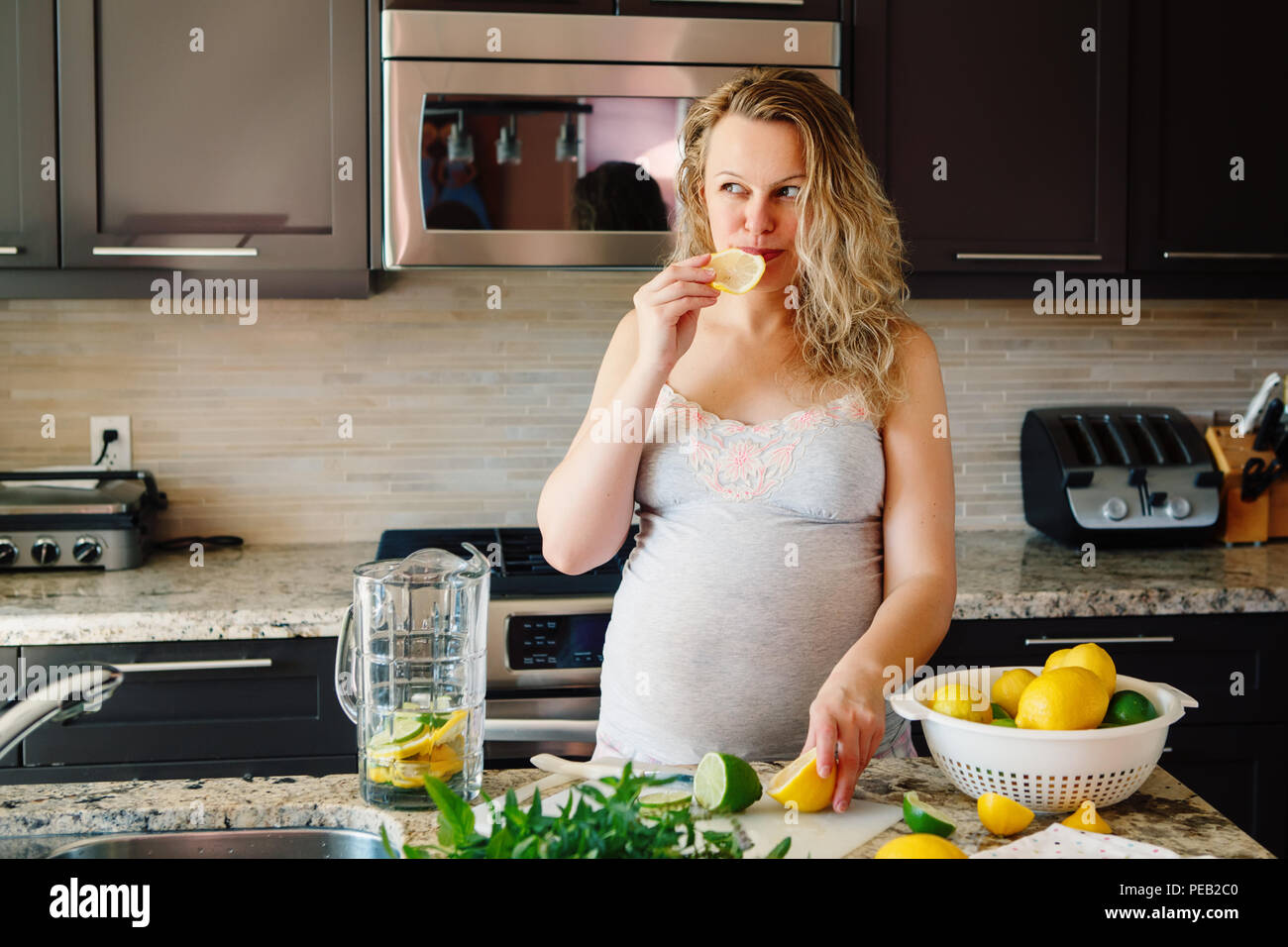 Portrait of white Caucasian blonde pregnant woman eating citrus lemon making juice standing in kitchen at home looking away, healthy lifestyle concept Stock Photo