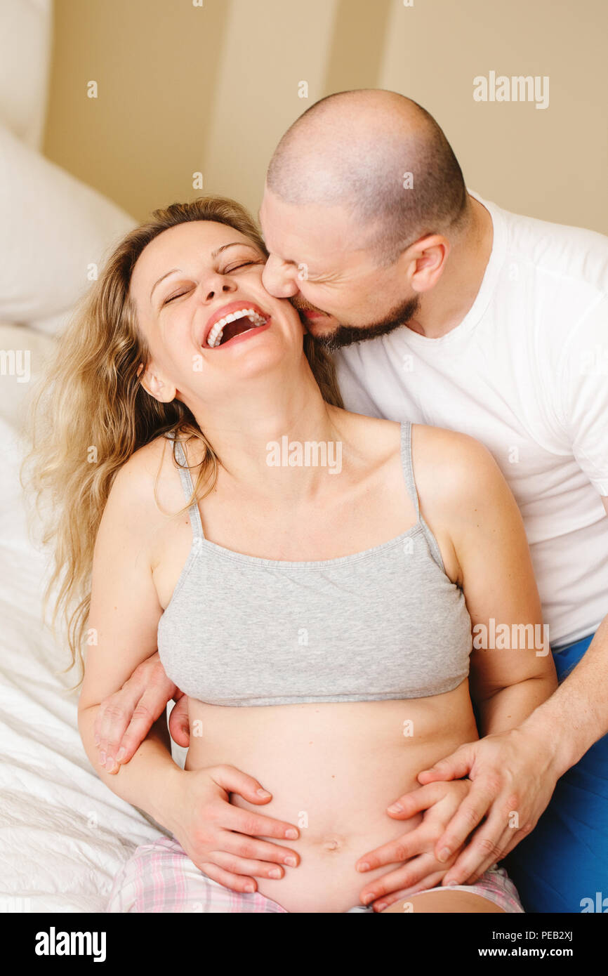 Portrait of smiling laughing white Caucasian young middle age couple, pregnant woman with husband in room on couch hugging cuddling, lifestyle materni Stock Photo