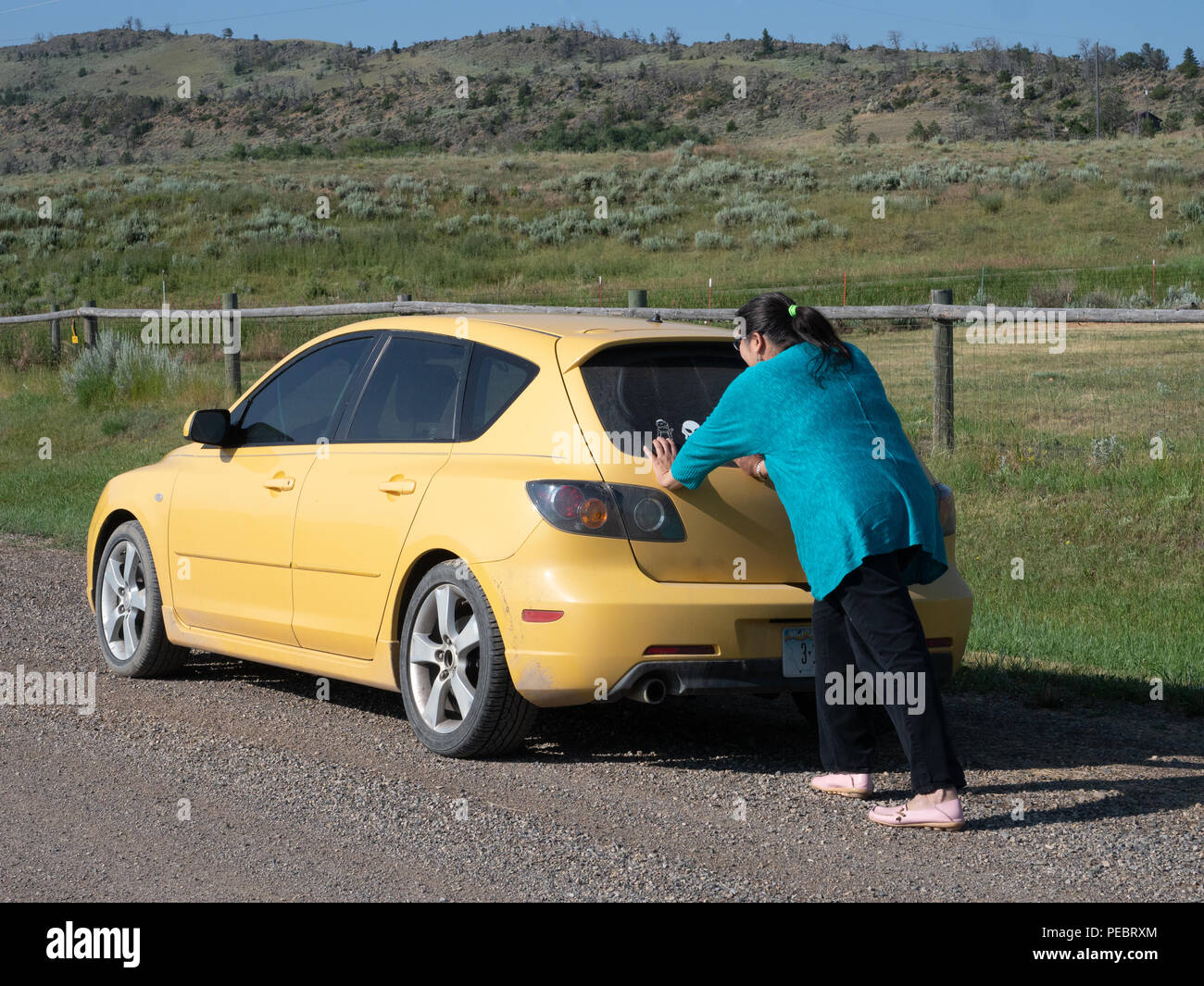 Hispanic woman pushing a compact yellow car after it has broken down in a rural area. Stock Photo