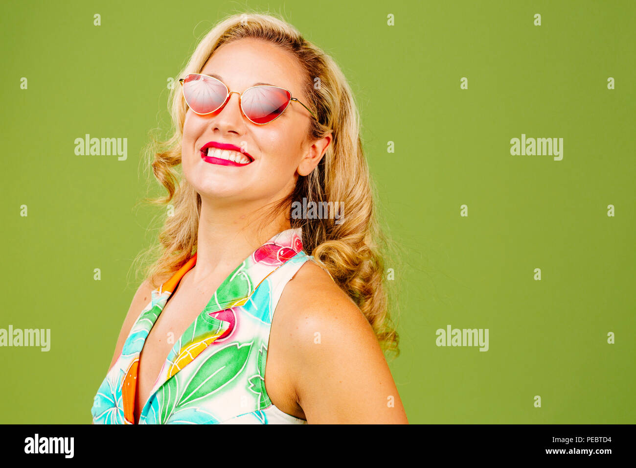 Smiling blonde woman with red sunglasses and summer dress tilting her head back, isolated on green studio background Stock Photo