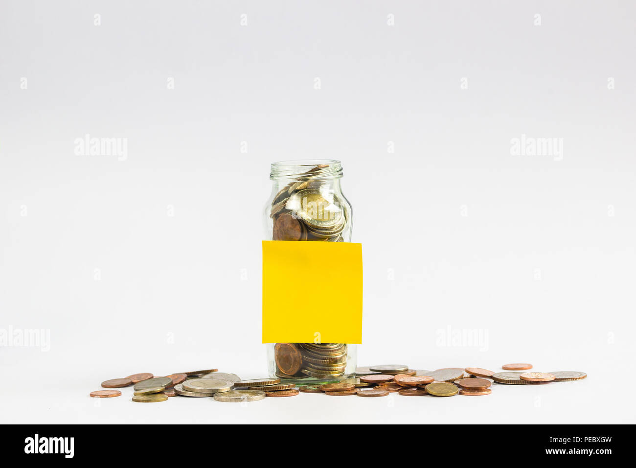 Savings, Investment growing money concept : A full coins in a clear glass jar with a empty yellow post paper and a coin on the floor on white backgrou Stock Photo