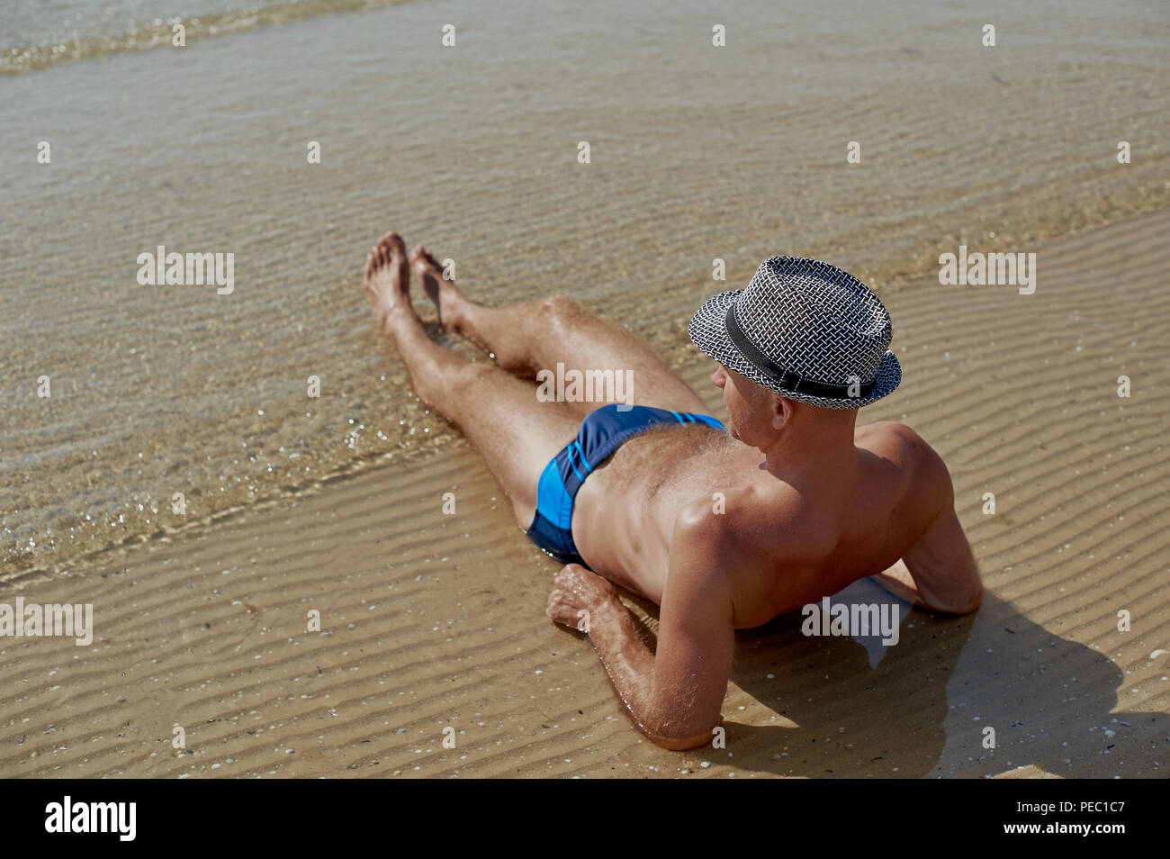 Summer lifestyle portrait of pretty young suntanned man in a hat. Enjoying life and sitting on the beach, time to travel. Looking at the sea Stock Photo
