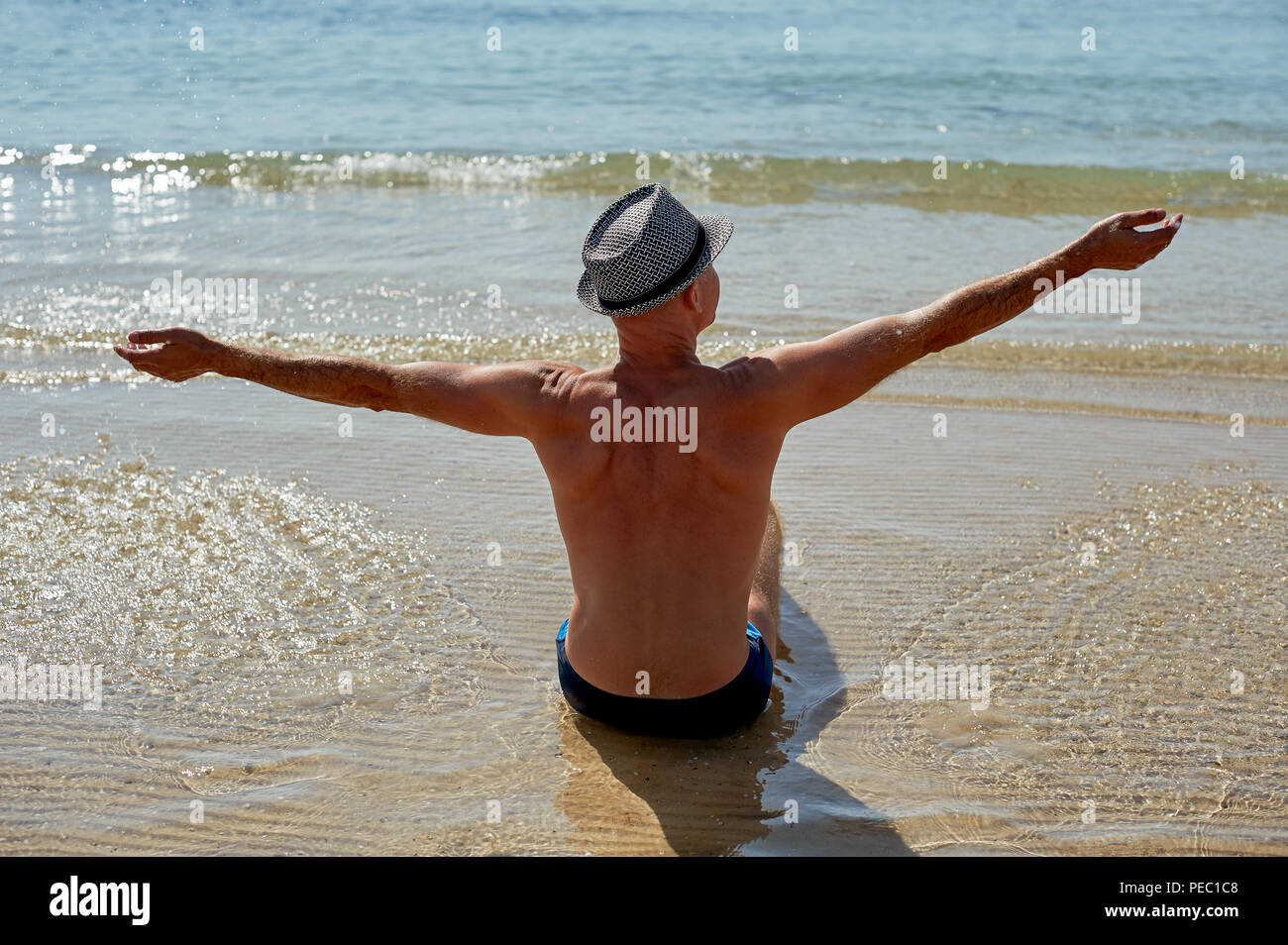 Summer lifestyle portrait of pretty young suntanned man in a hat. Enjoying life and sitting on the beach, time to travel. Looking at the sea Stock Photo