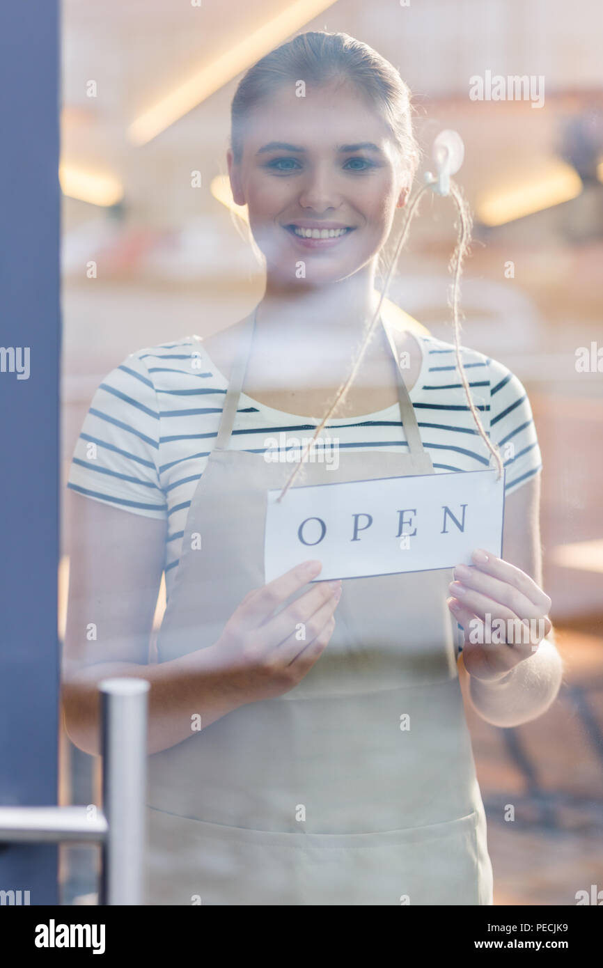In the morning. Cheerful nice positive woman standing behind the glass door and smiling to you while opening the cafe in the morning Stock Photo