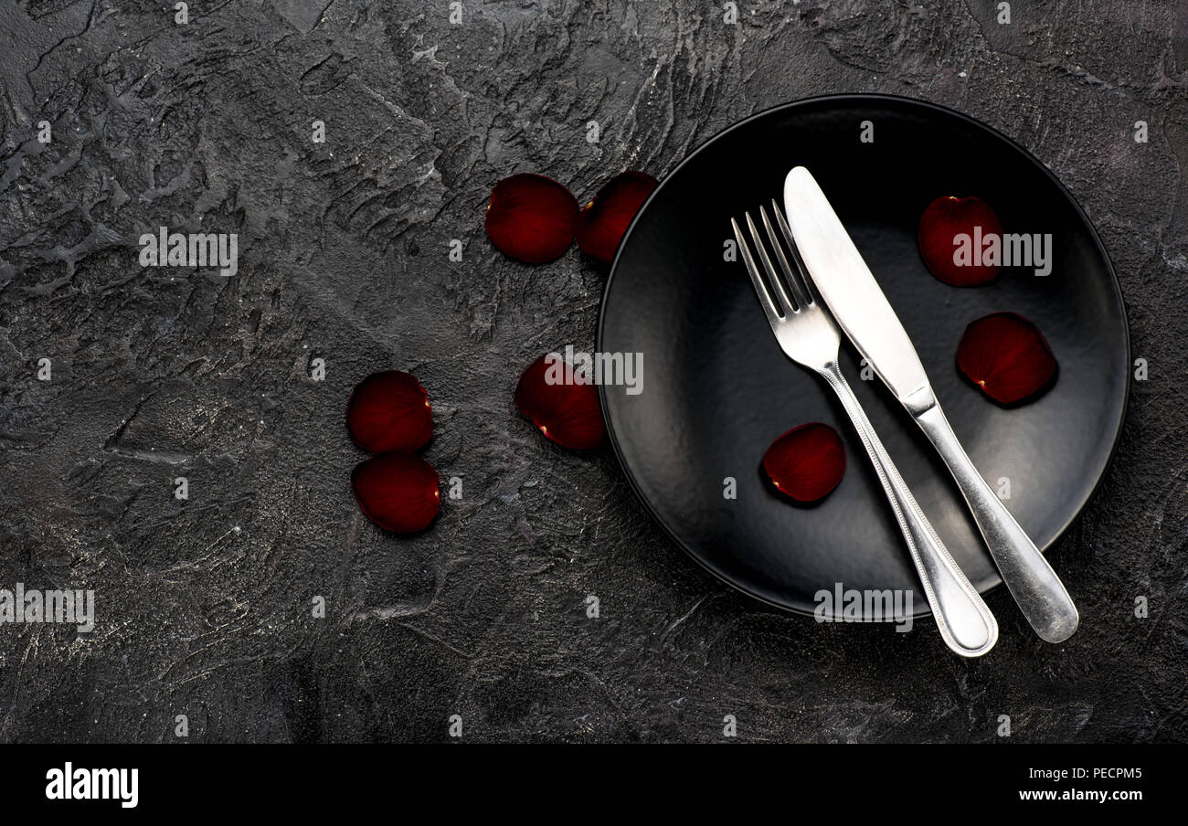 Fork, knife with red napkin, roses and black plate. On dark table