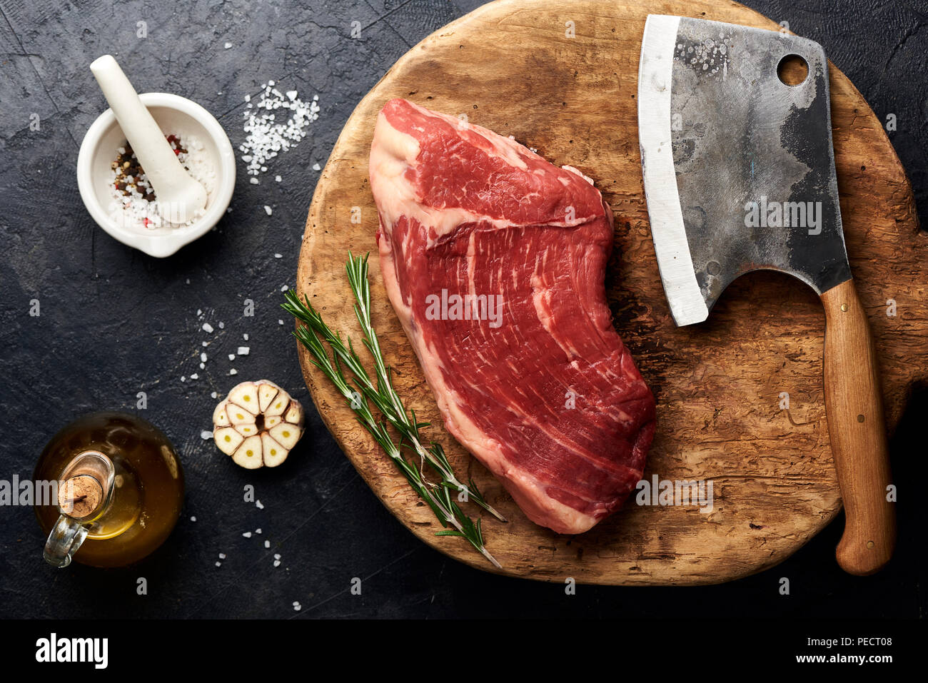 Raw fresh marbled meat Black Angus steak and meat cleaver on wooden board. Meat on black background with rosemary, spices, olive oil and garlic. Top view. Stock Photo