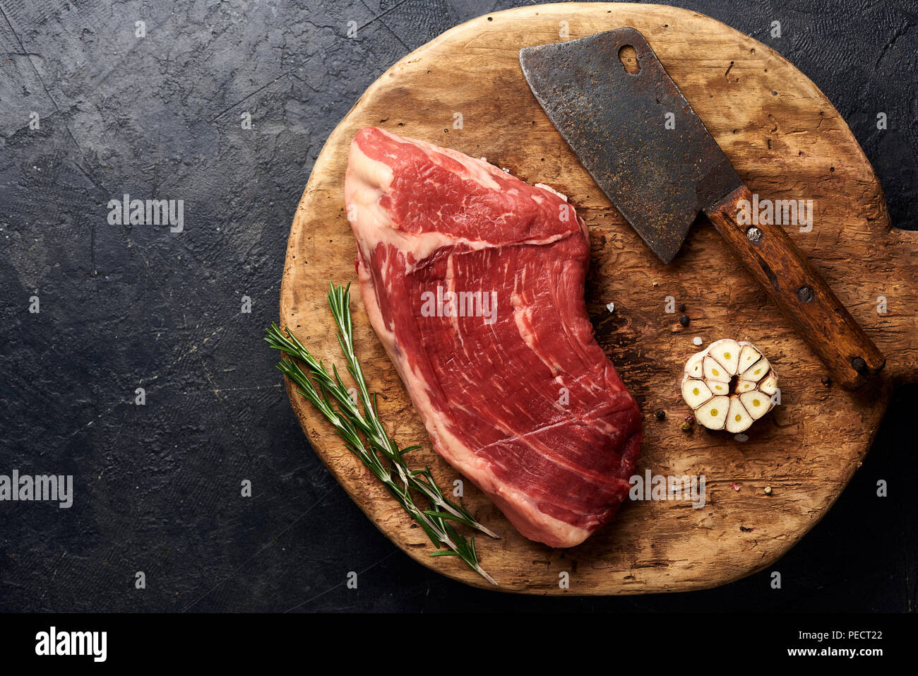 Raw fresh marbled meat Black Angus steak and vintage meat cleaver on wooden board. Meat on black background with rosemary and garlic. Copy space. Top view. Stock Photo