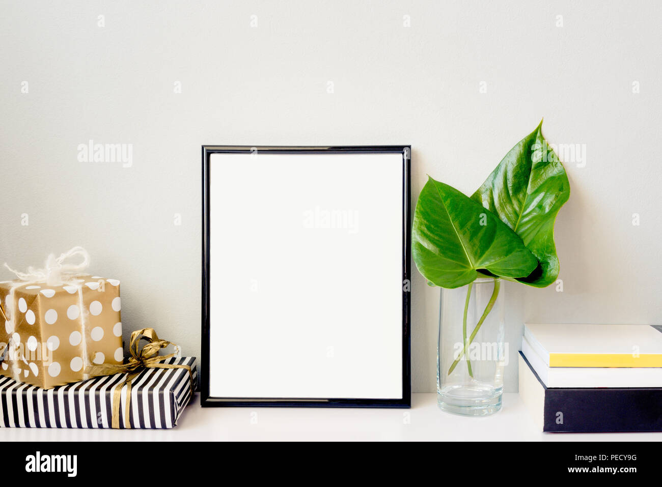 Black photo frame, green plant in a chrystal vase, gift boxes and a pile of books arranged against emty grey wall. Frame mock-up. Stock Photo