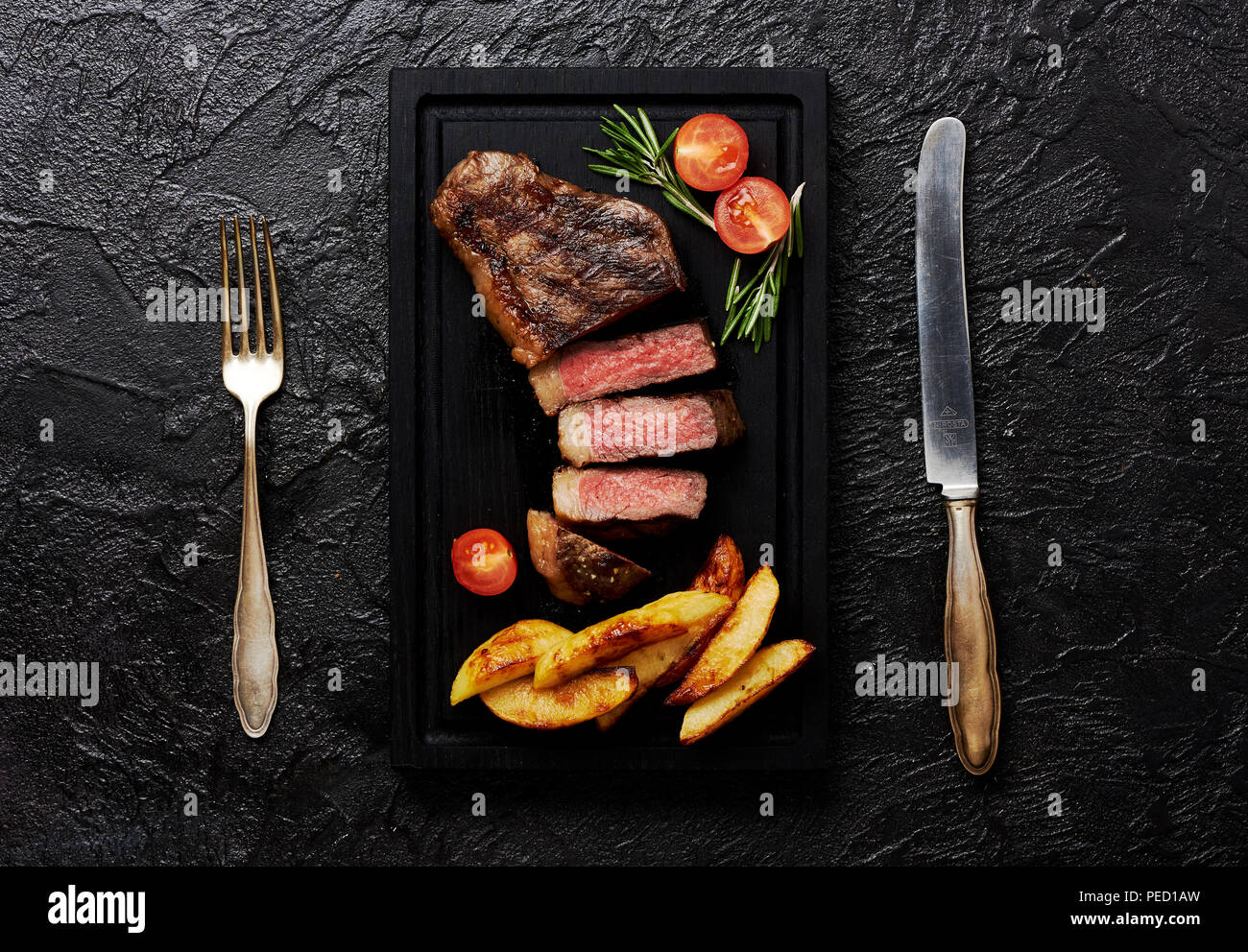 Meat Picanha steak, traditional Brazilian cut with tomatoes, potato wedges and rosemary on black meat cutting board. Steak with fork and knife. Black concrete background. Stock Photo