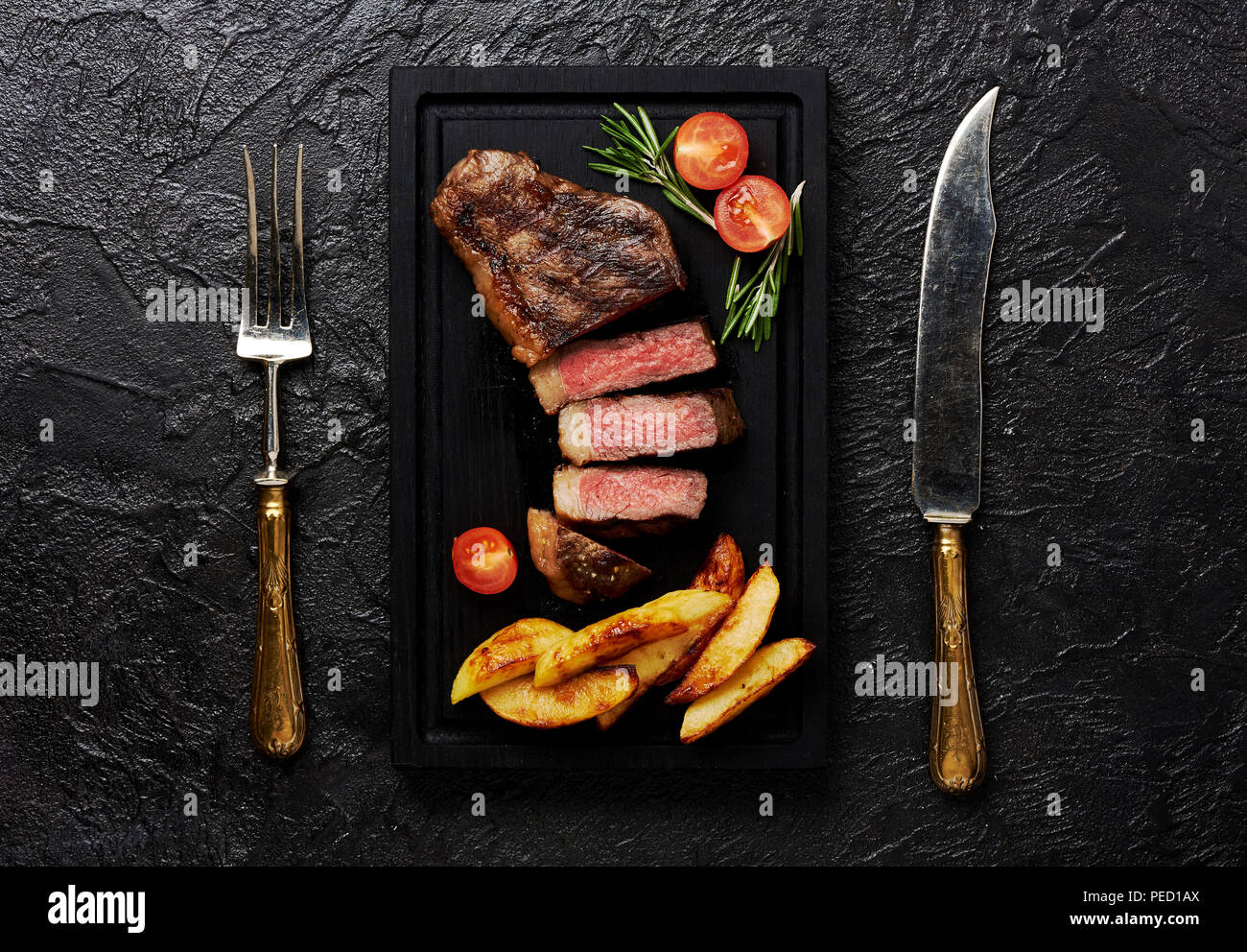 Meat Picanha steak, traditional Brazilian cut with tomatoes, potato wedges and rosemary on black meat cutting board. Steak with fork and knife. Black concrete background. Stock Photo