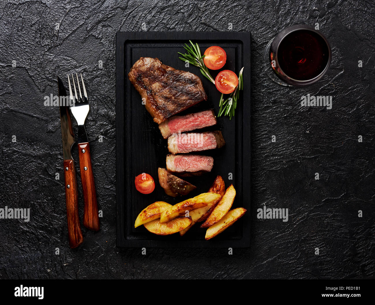 Meat Picanha steak, traditional Brazilian cut with tomatoes, potato wedges and rosemary on black meat cutting board. Steak and a glass of red wine with fork and knife. Black concrete background. Stock Photo