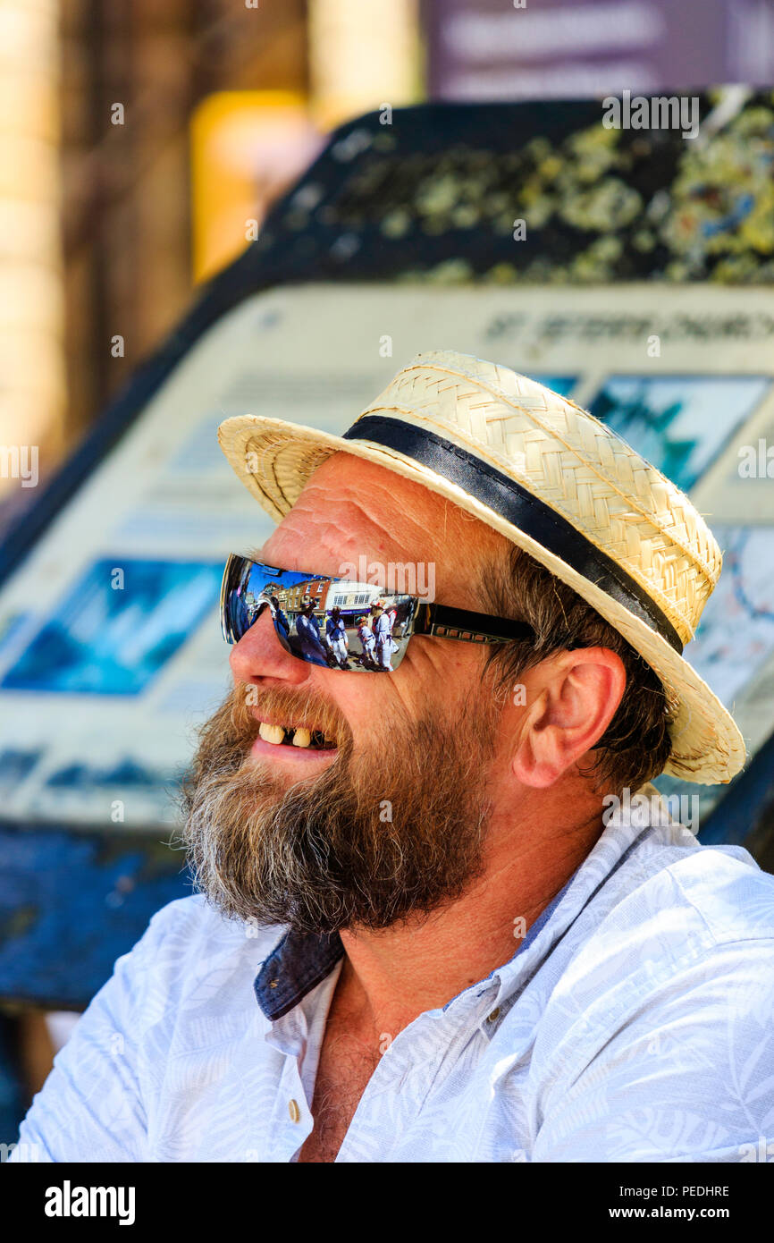 Mature caucasian man, face, side view. Wears straw boater, sunglasses with reflection of Morris men in. Dark beard, mouth open, some missing teeth. Stock Photo