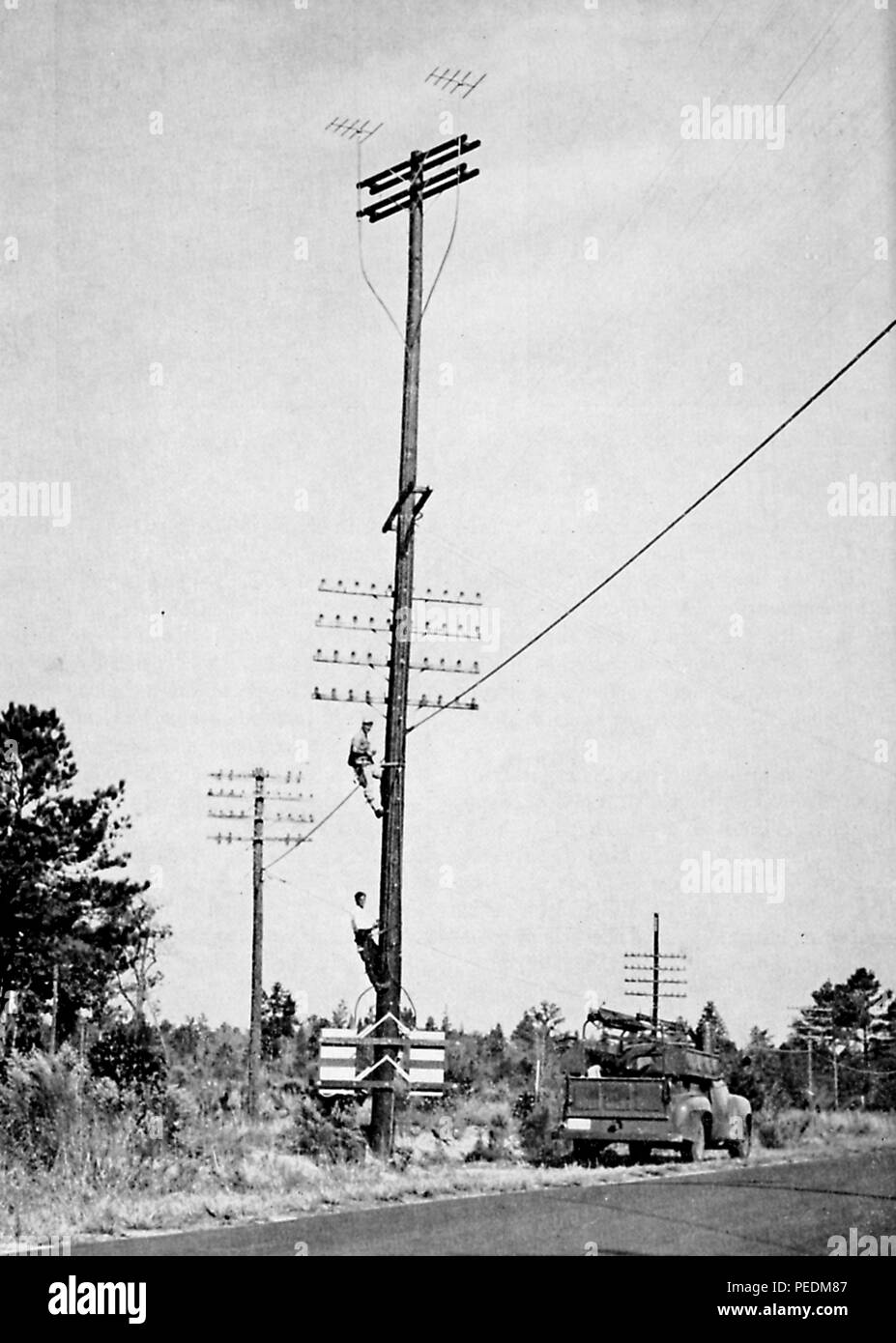Black and white photograph showing two men scaling a multiple transmission type telephone pole with a truck and trees in the background, from volume XXXI of the 'Bell Telephone Magazine, ' published in New York by the American Telephone and Telegraph Co, 1922. Courtesy Internet Archive. () Stock Photo