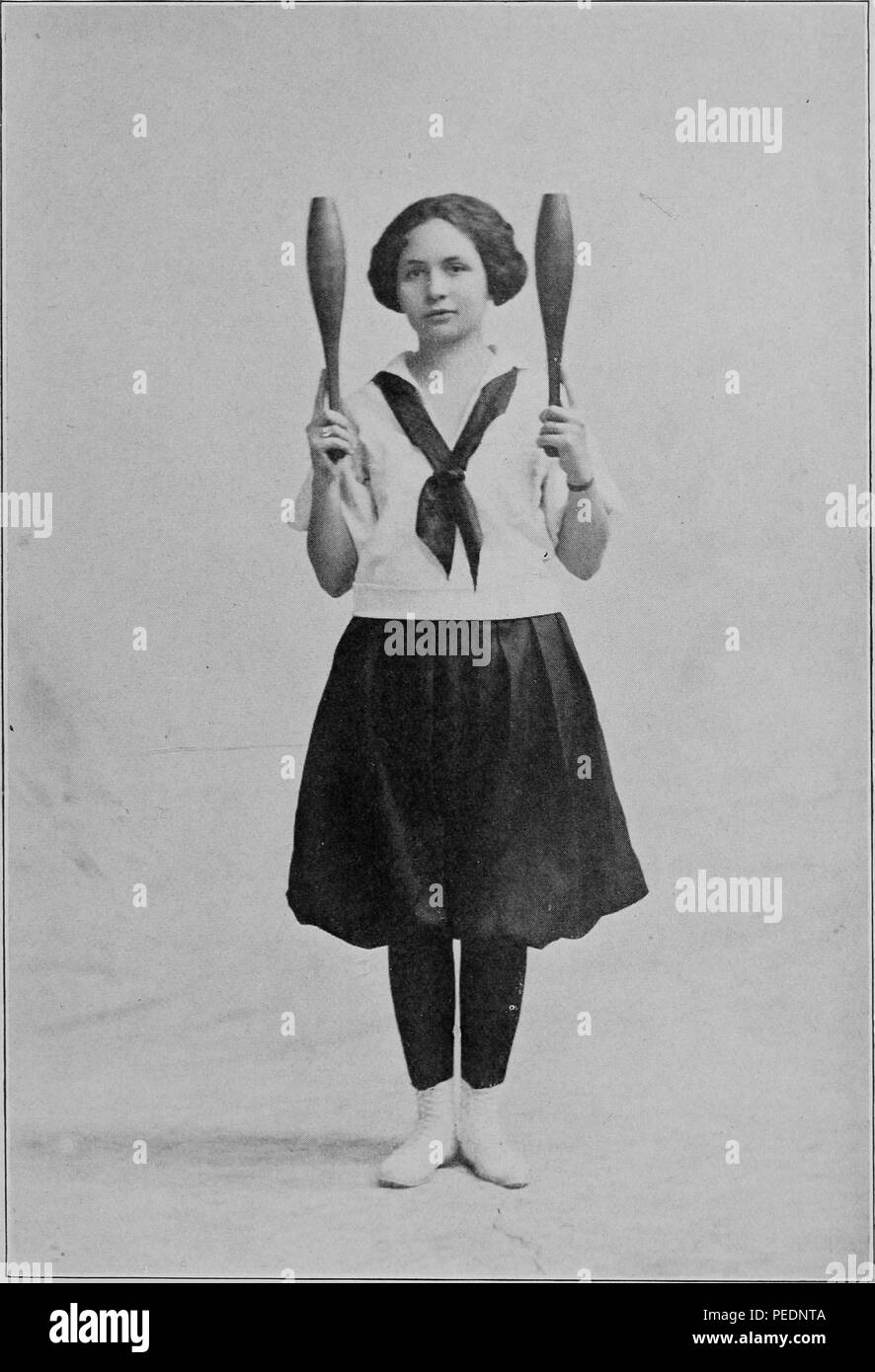 Black and white full-length photograph of a schoolgirl, facing the viewer, with a neutral expression, wearing Victorian women's fitness attire, and holding two clubs vertically in order to illustrate the fundamental position for carrying Indian Clubs, 1922. Courtesy Internet Archive. () Stock Photo