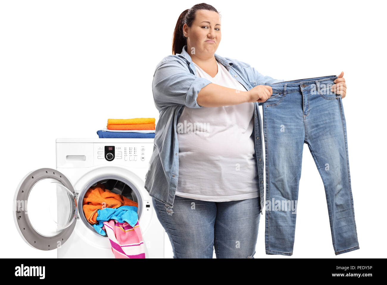 Disappointed young woman holding a pair of shrunken jeans with a washing machine behind her isolated on white background Stock Photo