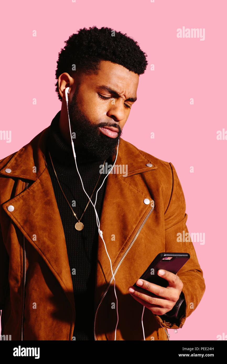 Portrait of a cool man with beard and headphones looking at phone with a frowning reaction, isolated on pink studio background Stock Photo