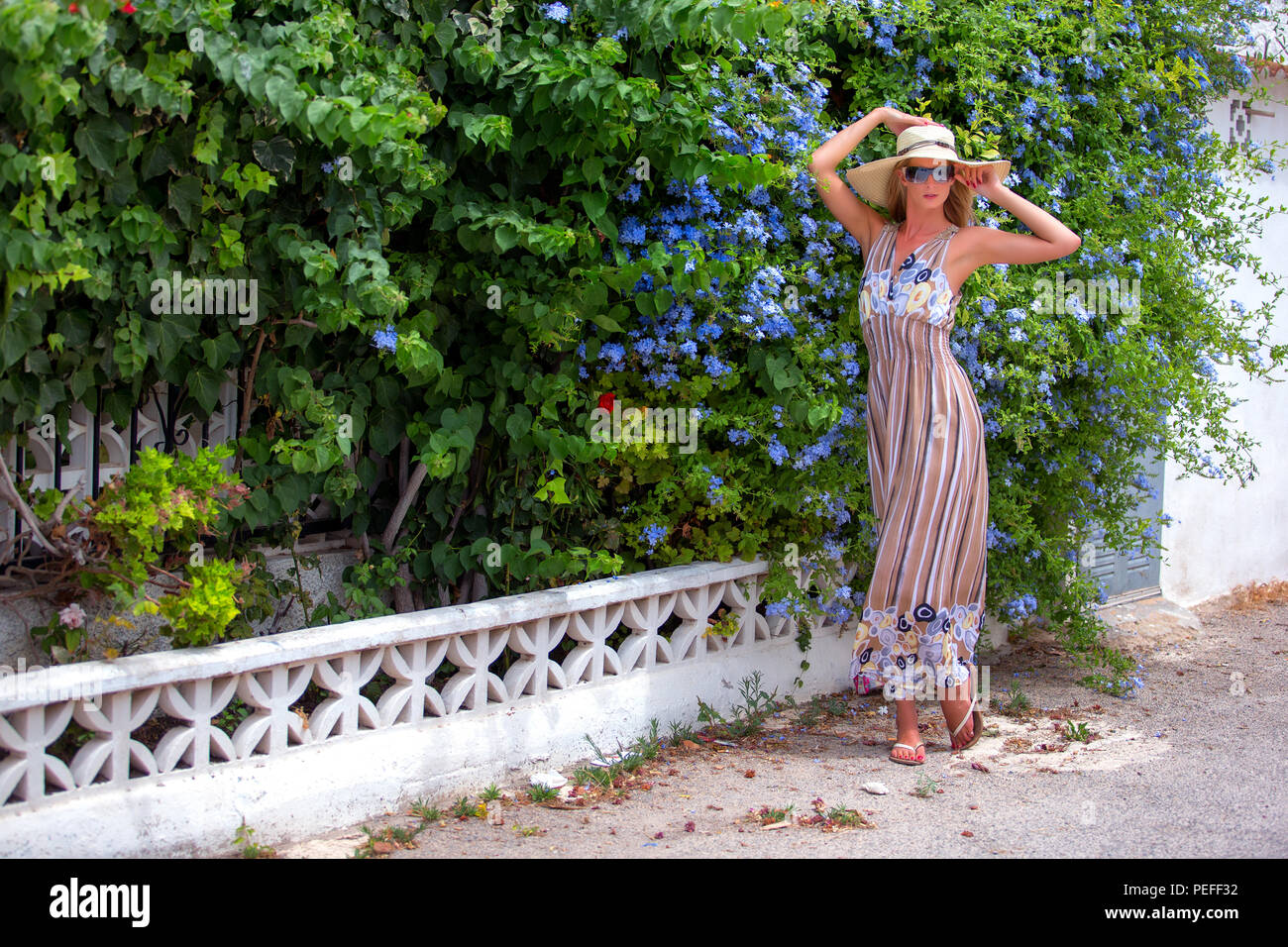 Tall blonde haired female standing on a pavement in front of a Spanish garden wearing a long stripey dress and a wide brimmed hat. Stock Photo