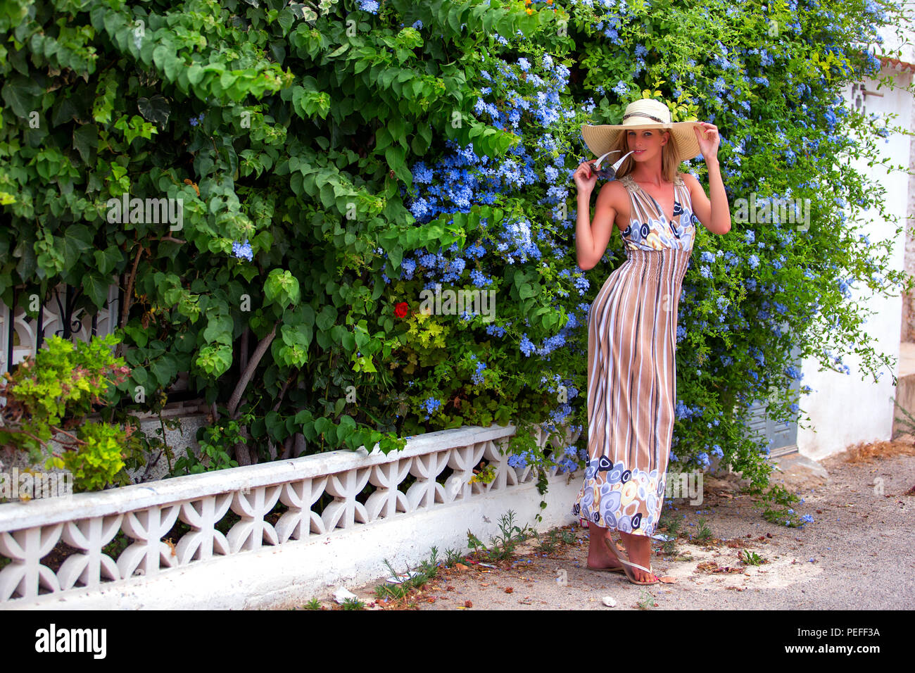 Tall blonde haired female standing on a pavement in front of a Spanish garden wearing a long stripey dress and a wide brimmed hat. Stock Photo
