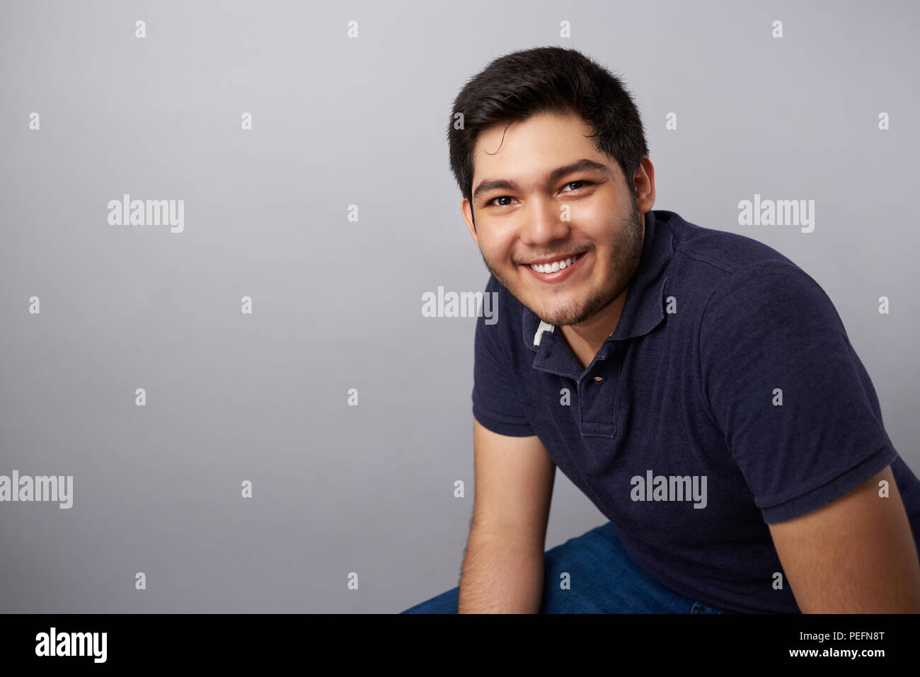 Cheerful smiling young hispanic man sit on gray studio background Stock Photo
