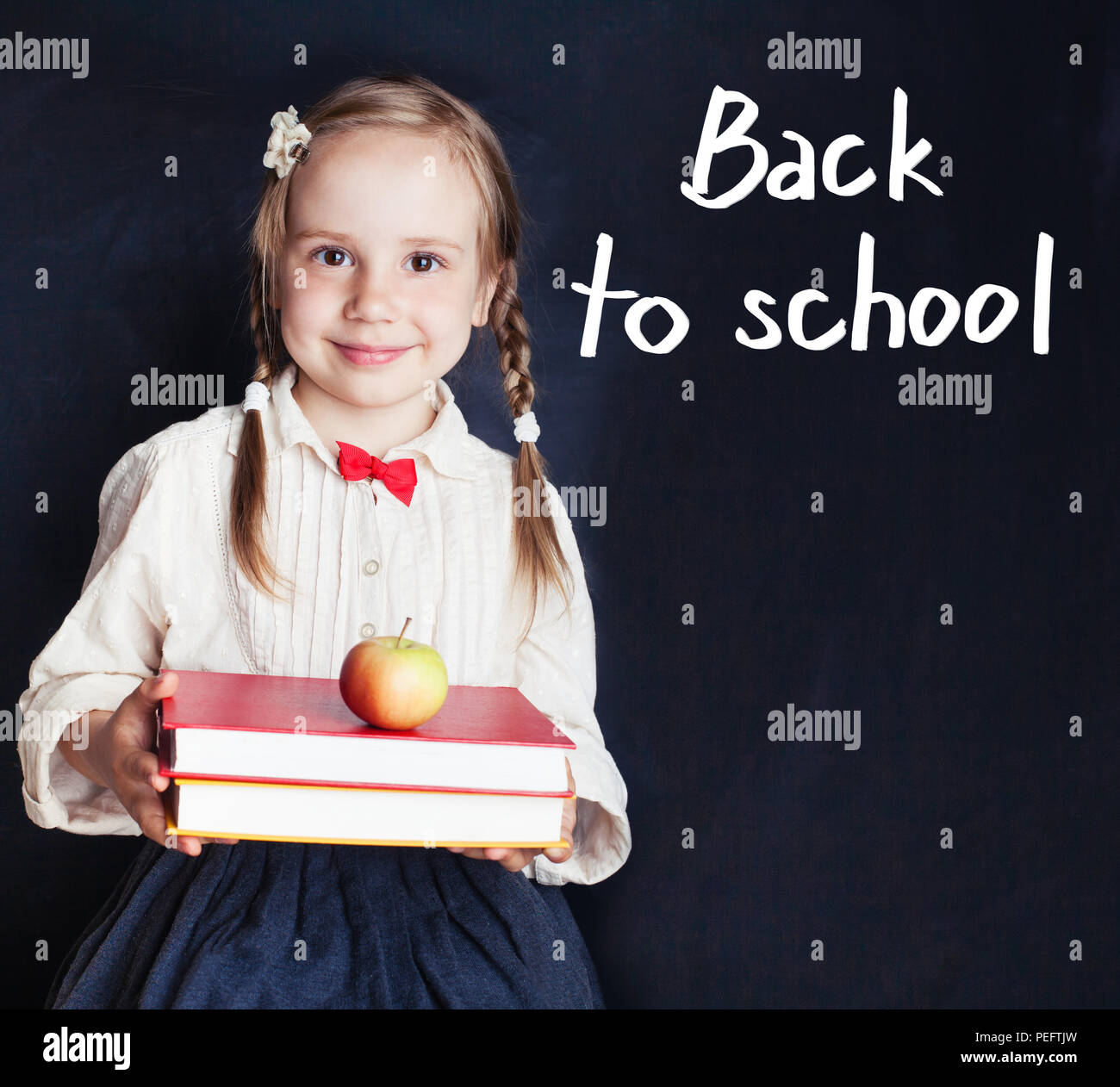 Little girl student in school uniform smiling and holding pile of books ...