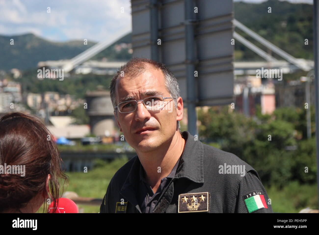 Genoa, Italy. 15th Aug, 2018. Fireman Emanuele Gissi answers questions from journalists. One day after the collapse of the highway bridge Ponte Morandi (background), it is still being searched for survivors in the enormous ruins. Credit: Fabian Nitschmann/dpa/Alamy Live News Stock Photo
