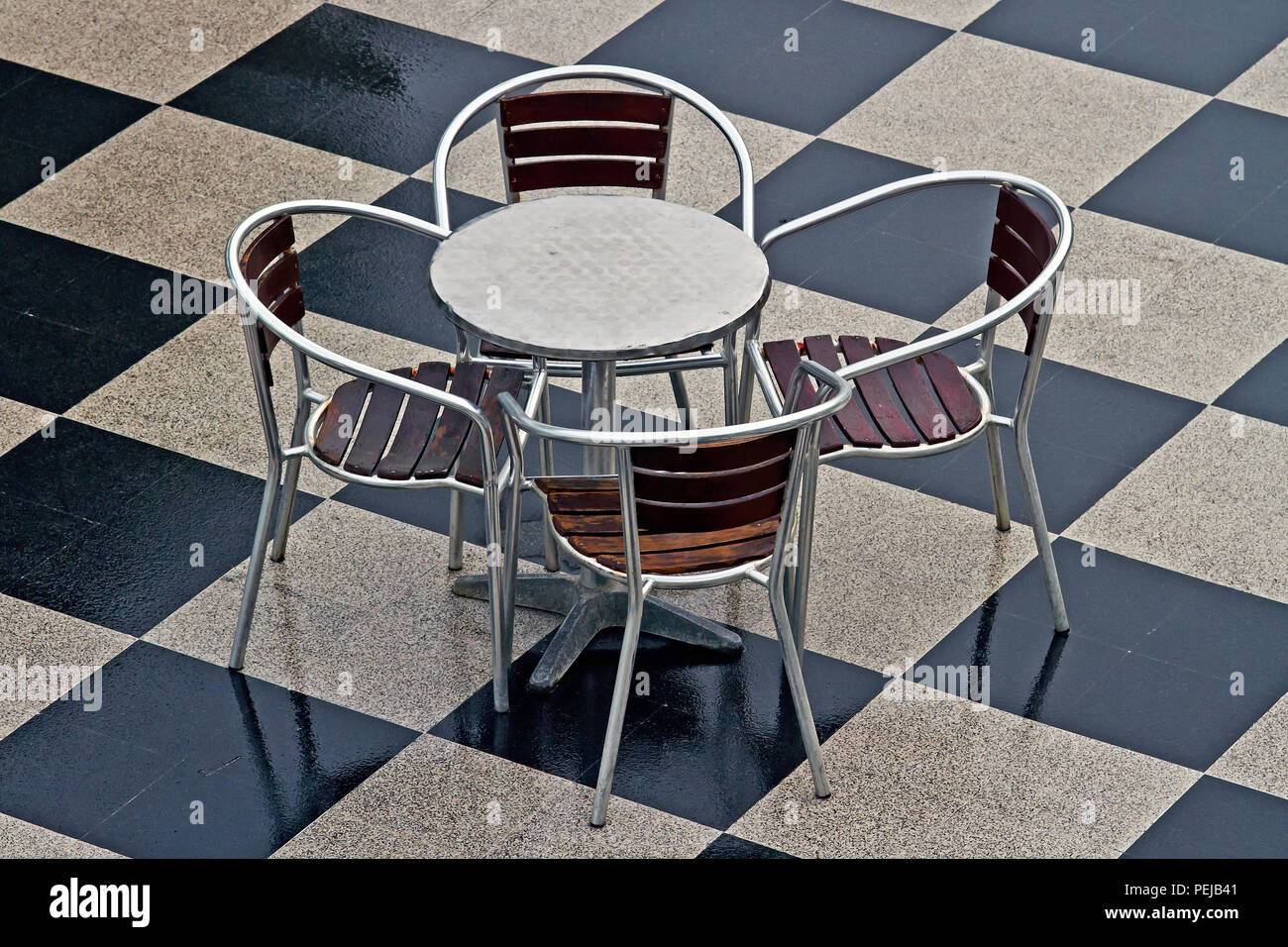 A vacant chrome table and four chairs sit unused on a checkerboard tile floor. Stock Photo