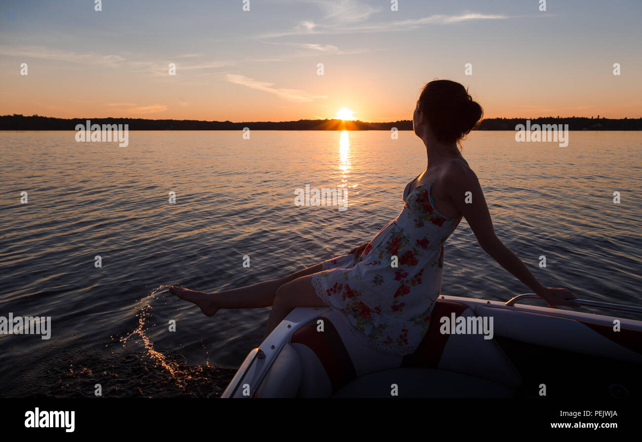 Young woman sitting on the edge of a boat on a lake at sunset. Stock Photo