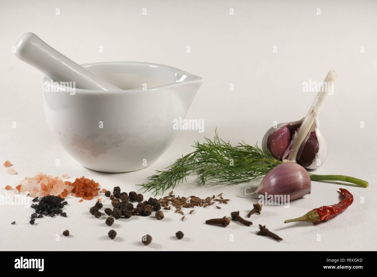 White ceramic mortar and pestle fresh dill garlic cumin seeds Himalayan pink salt Hawaii red aloes salt Hawaii black pearl salt cloves chili pepper bl Stock Photo