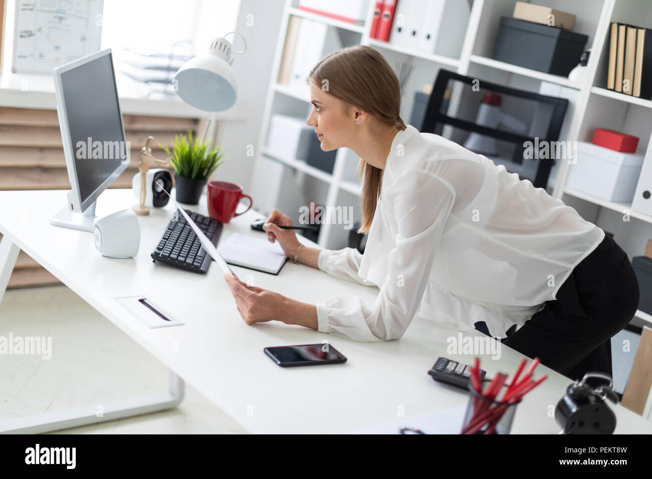 A young girl stands bent near a table in the office. The girl works with a  computer, notepad and documents Stock Photo - Alamy