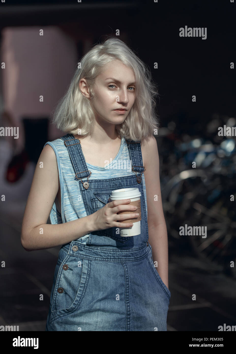 Portrait of beautiful Caucasian teenage young blonde alternative model girl woman in blue tshirt, jeans romper looking in camera holding cup of coffee Stock Photo